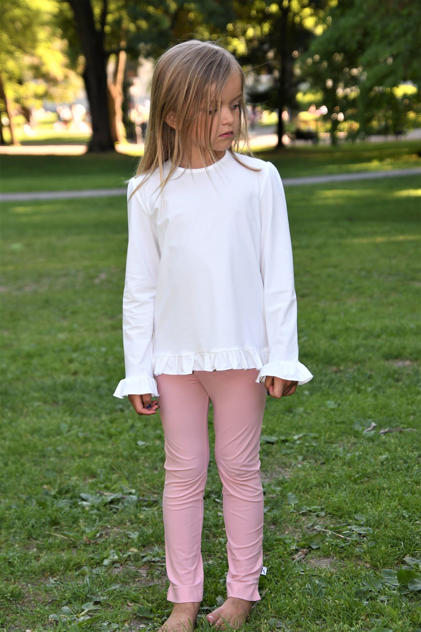 Young girl in a white blouse and pink pants standing in a grassy area with trees in the background