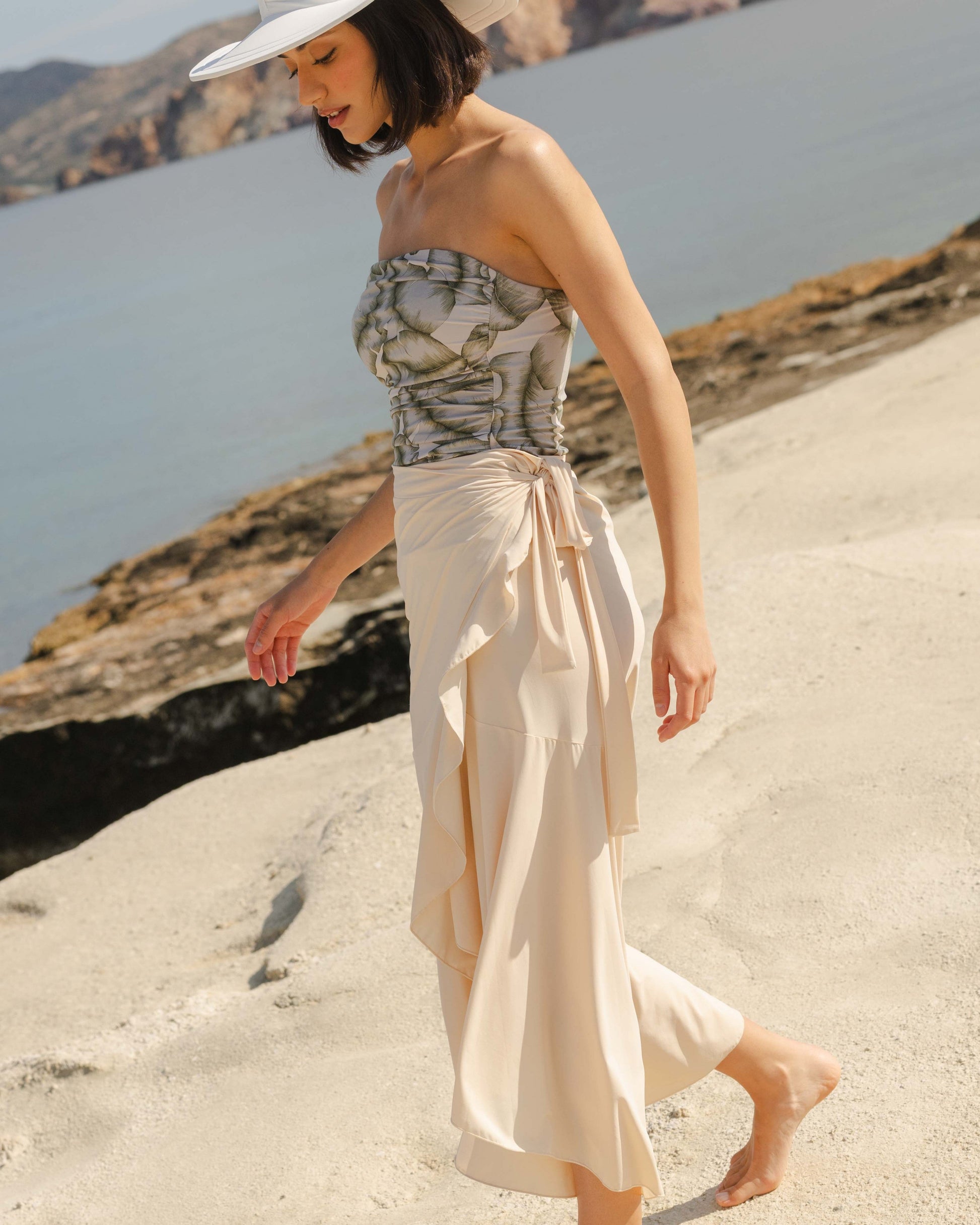 Woman in a strapless swimsuit and wide-brimmed hat standing on a sandy beach with mountains in the background.